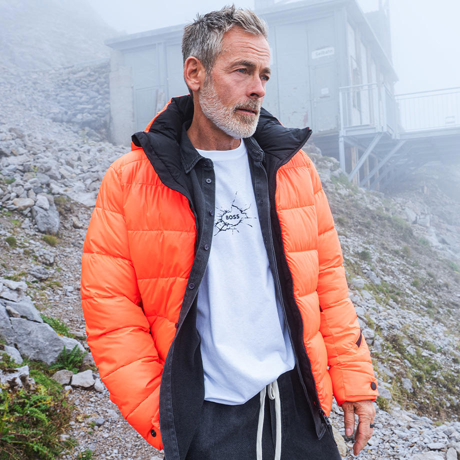 Ein Mann steht in nebliger Berglandschaft und trägt eine auffällige, orangefarbene Steppjacke über einem weißen T-Shirt mit BOSS-Logo und einem offenen Hemd. Das moderne, sportliche Outfit kombiniert Funktionalität mit urbanem Stil – ideal für kühle Outdoor-Tage.