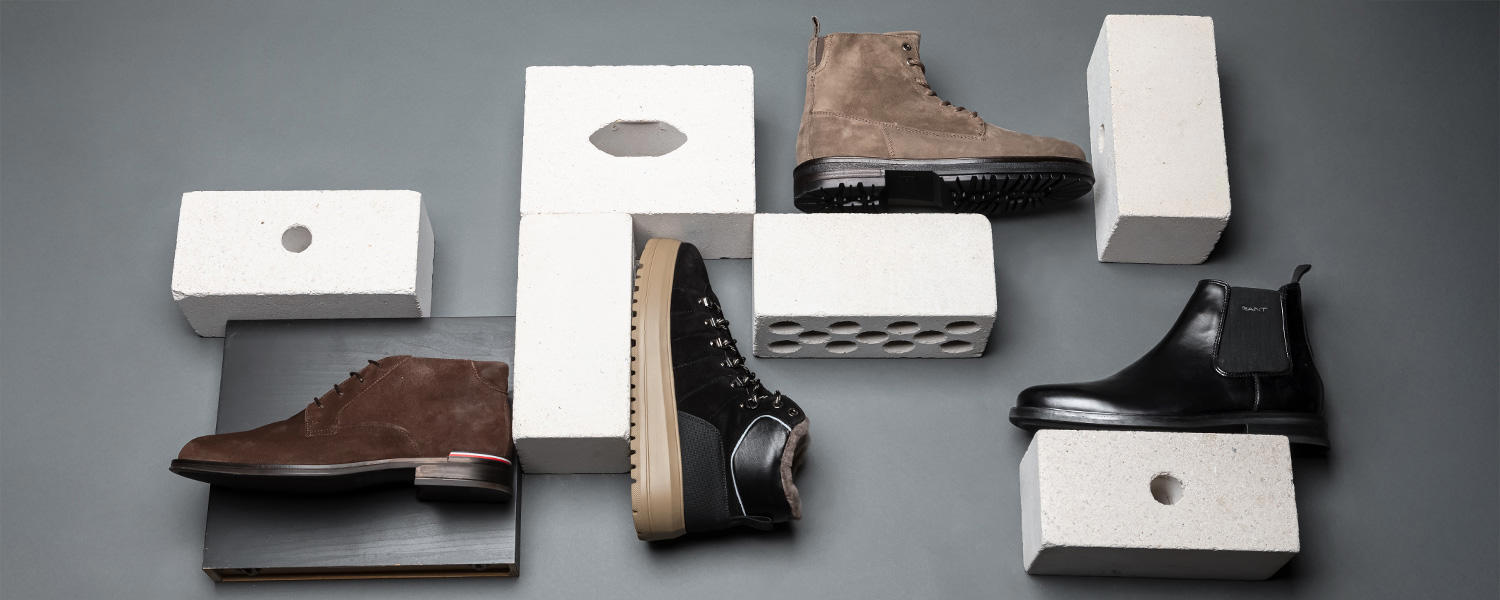 Verschiedene Herren-Stiefel und Boots aus Leder und Veloursleder auf grauem Hintergrund mit weißen Backsteinen dekoriert. Verschiedene Herren-Stiefel und Boots aus Leder und Veloursleder auf grauem Hintergrund mit weißen Backsteinen drapiert.
