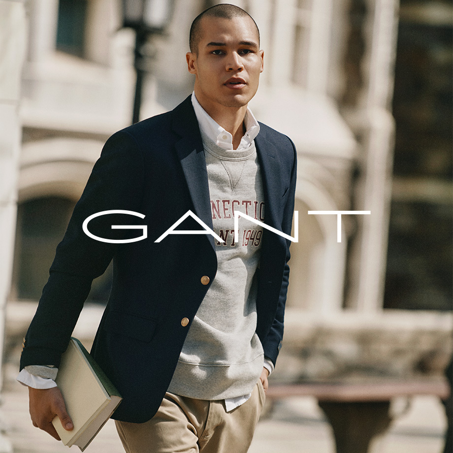 Mann in dunkelblauem Blazer mit grauem Sweatshirt, weißem Hemd und Chinohose von Gant, Buch in der Hand, draußen vor Gebäude.