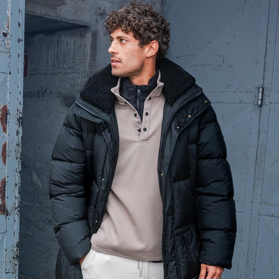 Ein Mann steht vor einer grauen Wand und trägt einen schwarzen Parka über einem hellbeigen Troyer und einer schwarzen Fleecejacke. Die Kleidung wirkt funktional und stylisch zugleich – perfekt für kühles Wetter. Die Szenerie vermittelt eine schlichte, urbane Winterstimmung.