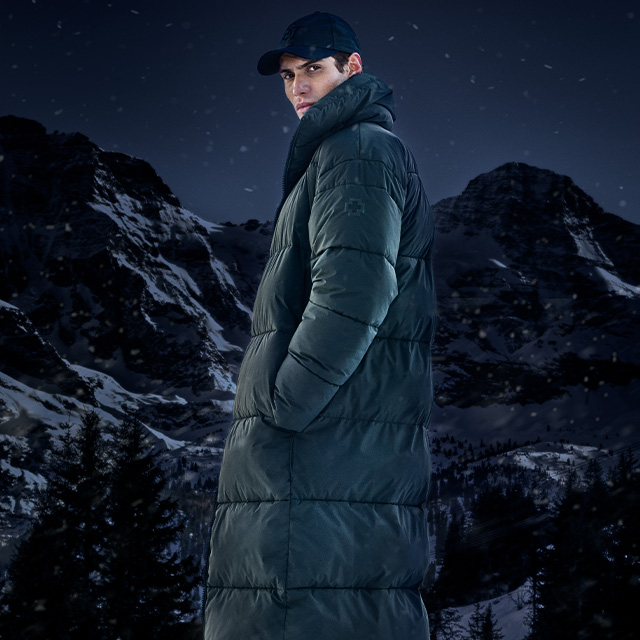 Mann in langem dunkelgrünen Daunenmantel und Baseballcap, draußen im Schnee vor Bergkulisse