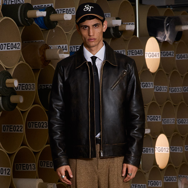 Mann in schwarzer Lederjacke mit weißem Hemd, Krawatte und Baseballcap, stehend vor Papierrollen