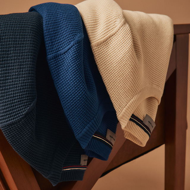 Drei Strickpullover in Dunkelblau, Royalblau und Beige, gefaltet über Stuhllehne gelegt