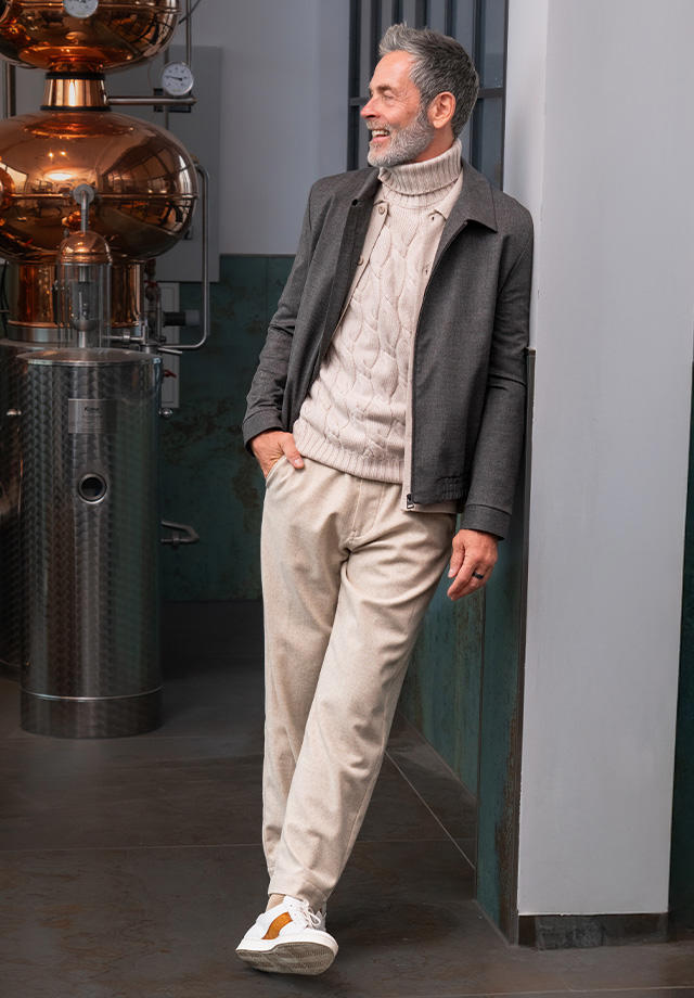 Älterer Mann in grauer Jacke mit beigem Zopfmuster-Rollkragenpullover und heller Hose, sportlich mit Sneakers