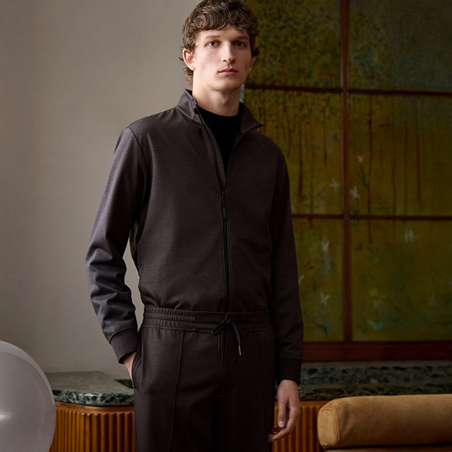 Mann in dunkelbraunem Freizeitanzug mit Stehkragen und Reißverschlussjacke, stehend im Wohnzimmer