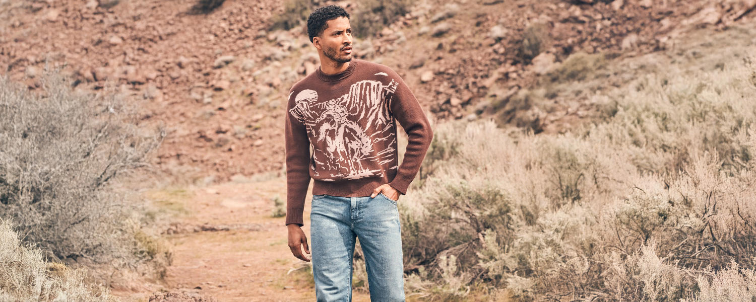 Ein authentisches Markenmotiv von Wrangler: Ein Mann in hellem Denim und braunem Pullover mit Western-Motiv steht inmitten einer kargen Landschaft – Sinnbild für Abenteuergeist, Freiheit und amerikanische Heritage-Mode.