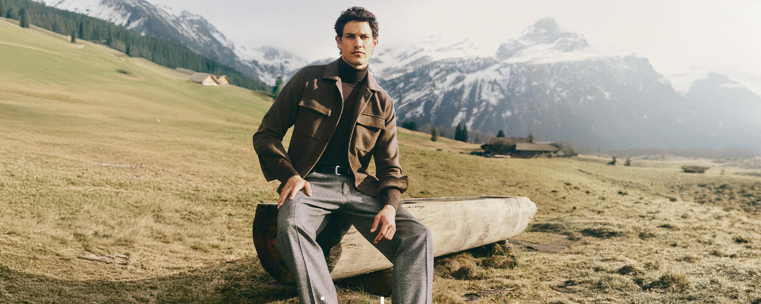 Ein elegantes Markenmotiv von Windsor: Ein Mann sitzt vor alpiner Bergkulisse auf einem Holzstamm und trägt einen braunen Overshirt-Jacket über einem dunklen Rollkragenpullover und grauen Wollhosen – eine Kombination aus stilvoller Modernität und klassischer Eleganz.