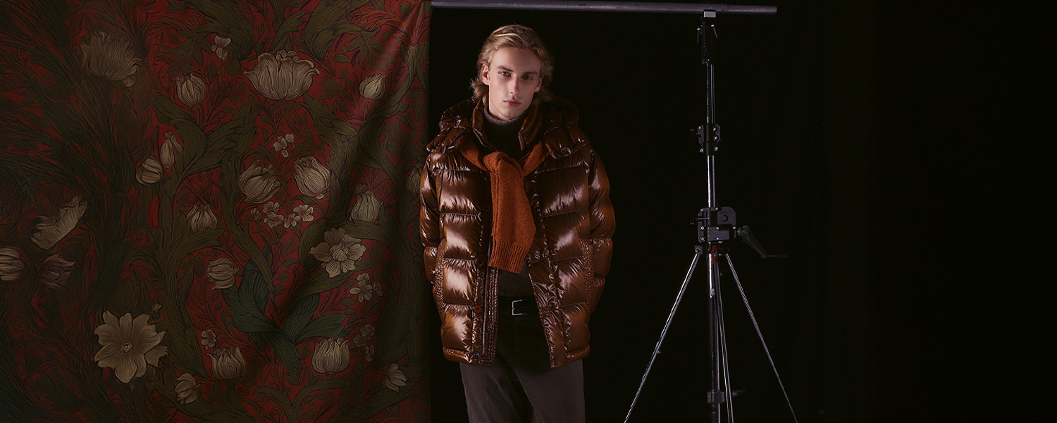 Mann trägt eine glänzende braune Daunenjacke von Peuterey, kombiniert mit dunkler Hose und orangefarbenem Pullover, der locker um den Hals gebunden ist, vor einem dunklen Hintergrund mit floralem Vorhang.