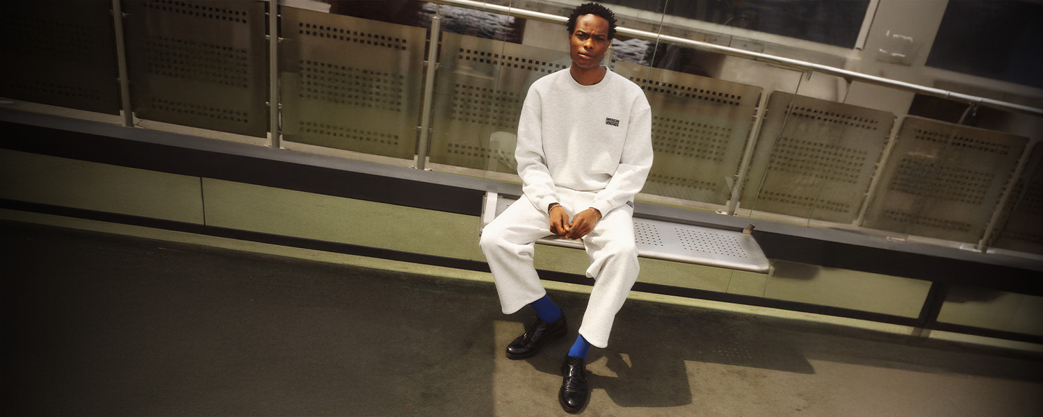 Mann in hellgrauem Sweatshirt und Sweathose von American Vintage, kombiniert mit schwarzen Lederschuhen und blauen Socken, sitzend auf Bank.