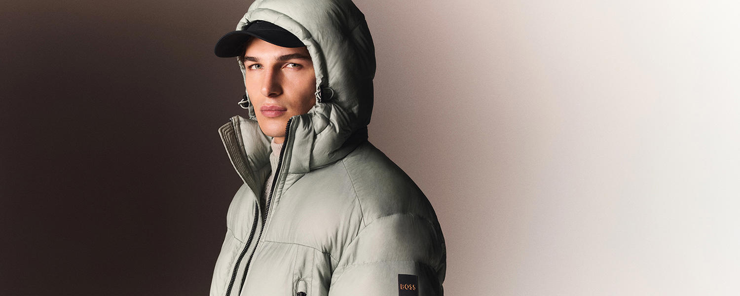 Mann in hellgrauer Daunenjacke mit Kapuze von Boss Orange, kombiniert mit schwarzer Baseballcap vor neutralem Hintergrund.