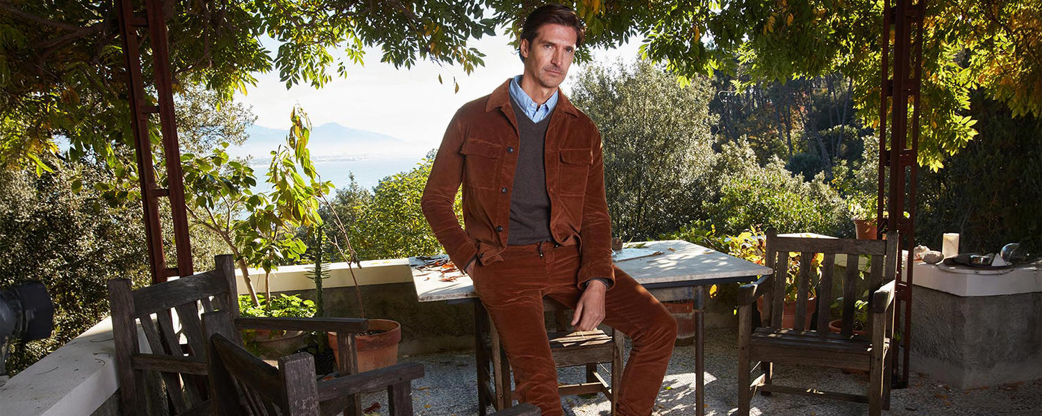 Mann in braunem Cord-Outfit von Masons, kombiniert mit Hemd und Pullover, sitzt auf einer Terrasse im Grünen mit Blick auf die Landschaft.