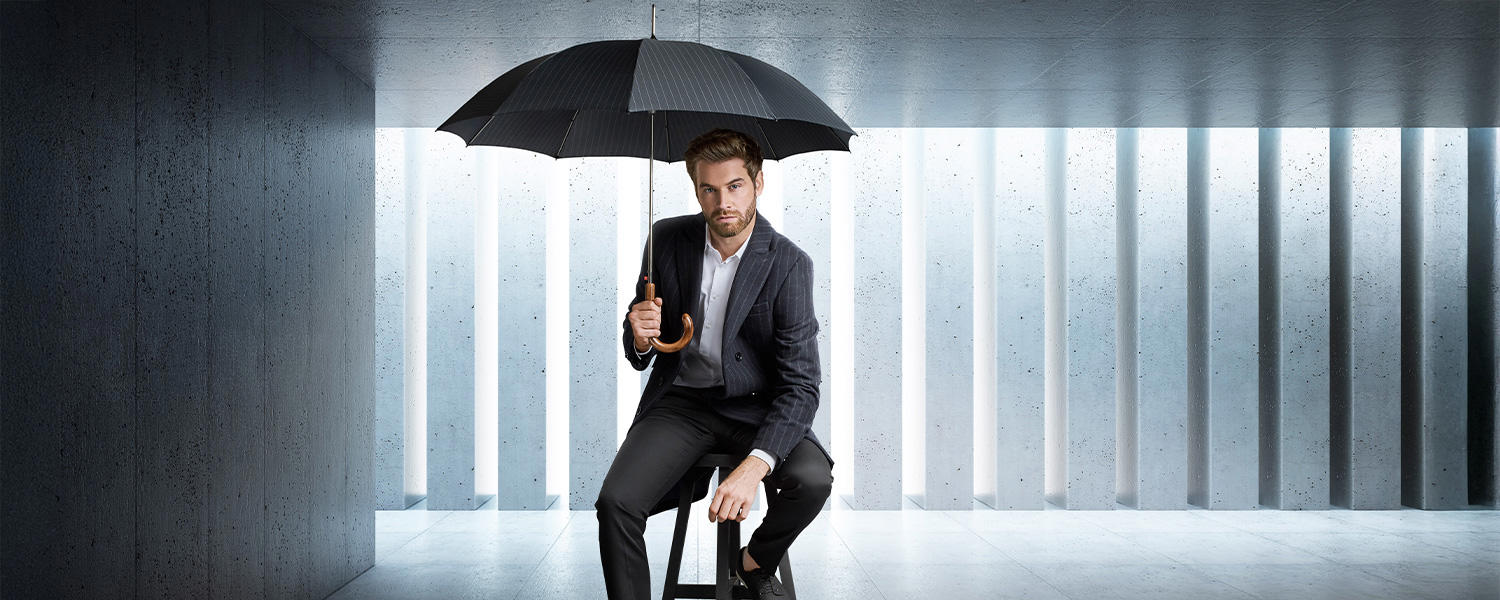 Mann im dunklen Anzug sitzt auf einem Hocker und hält einen schwarzen Regenschirm von Knirps in modernem, minimalistischem Raum.