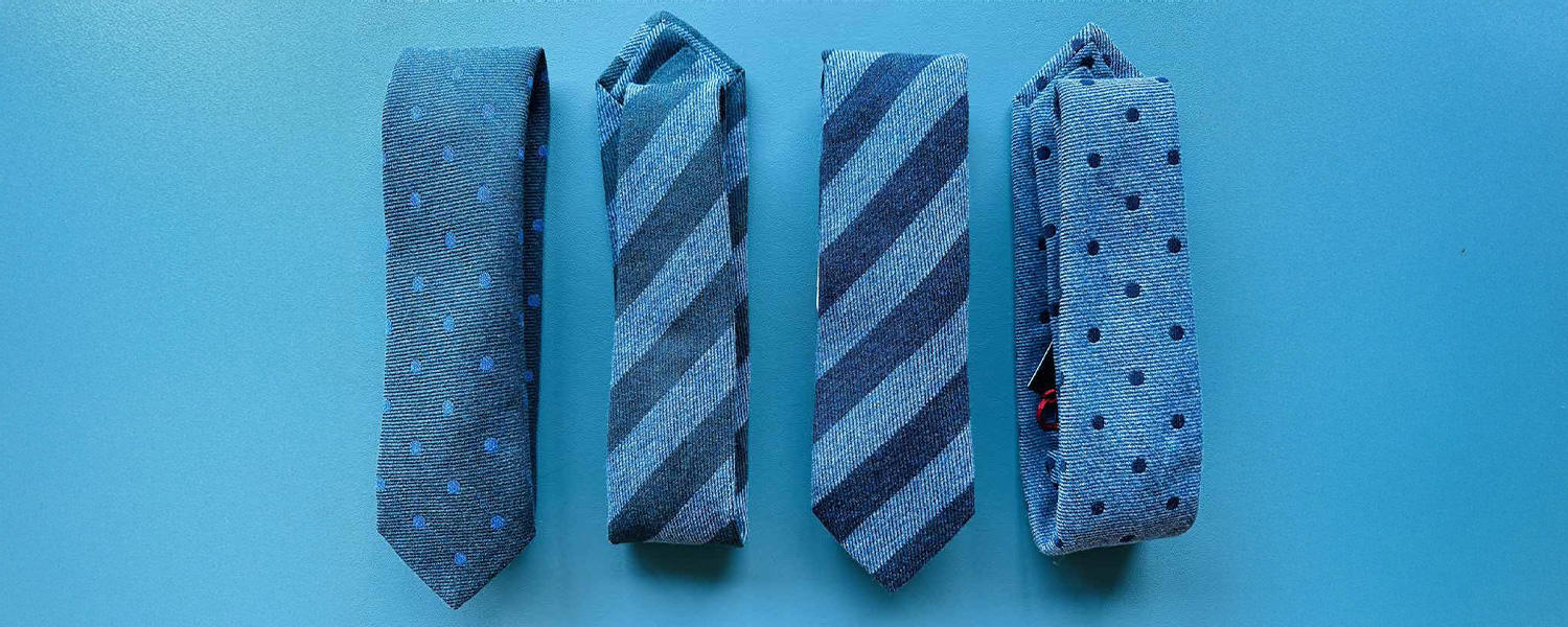 Vier blaue Krawatten von Ascot, zwei mit Punktmuster und zwei mit diagonalen Streifen, nebeneinander auf blauem Hintergrund platziert.