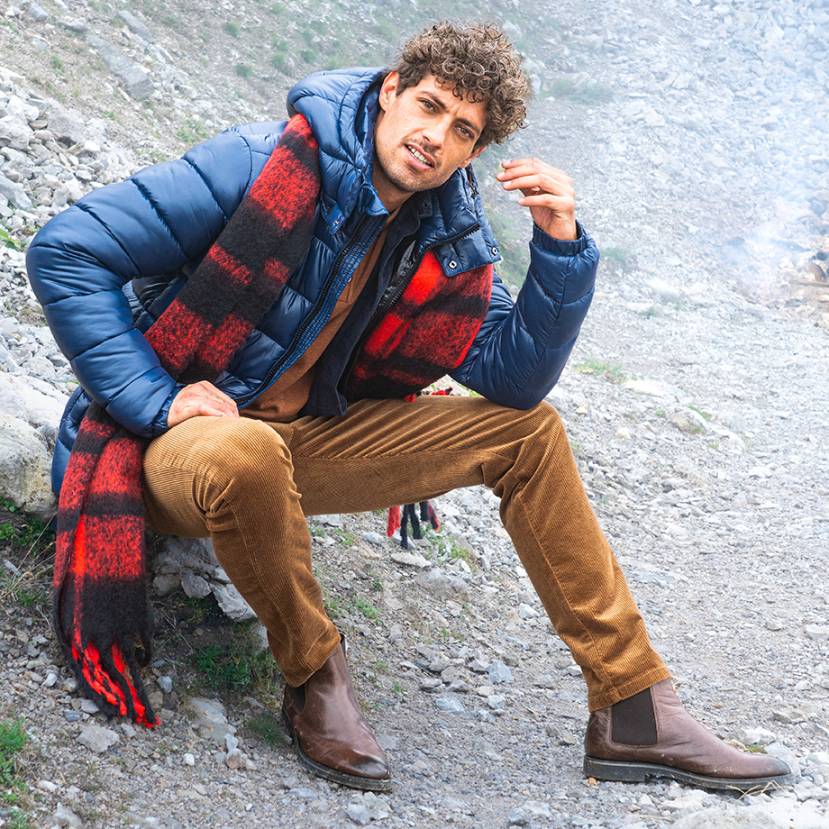 Auf dem Bild sitzt ein Mann in einer lässigen Herbst-/Winter-Kombination: blaue Steppjacke, braune Cordhose, braune Chelsea Boots und ein auffälliger, rot-schwarz karierter Schal. Im Hintergrund brennt ein kleines Feuer – ein Outdoor-Look mit rustikalem Flair.