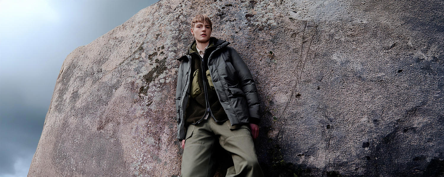 Ein ausdrucksstarkes Markenmotiv von Woolrich: Ein Mann lehnt lässig an einem Felsen und trägt eine dunkle Steppjacke kombiniert mit einer mehrlagigen Outdoor-Kombination in Grüntönen – perfekt für urbane Abenteuer und Naturerlebnisse.
