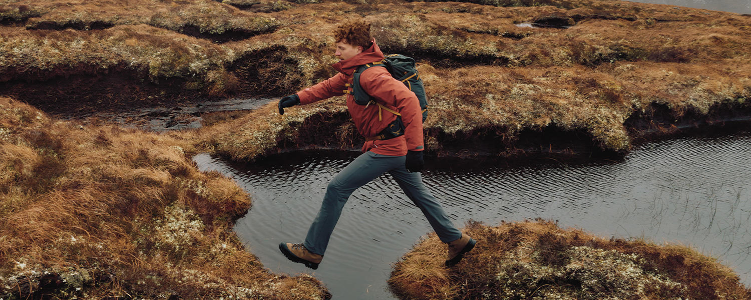 Ein Mann in funktionaler Schöffel Outdoor-Bekleidung – mit roter Jacke, Rucksack und Wanderschuhen – springt dynamisch über einen Wasserlauf inmitten einer herbstlichen Landschaft.
