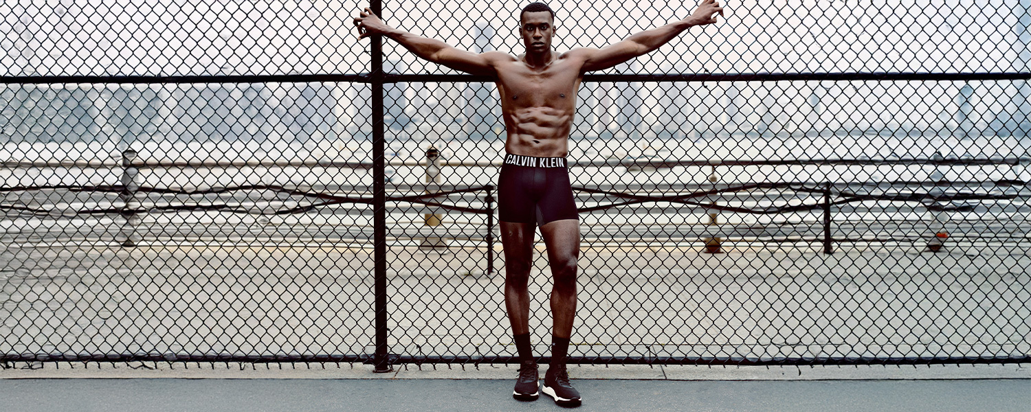Mann in schwarzer Calvin Klein Unterwäsche, stehend mit ausgebreiteten Armen vor Maschendrahtzaun im Freien.