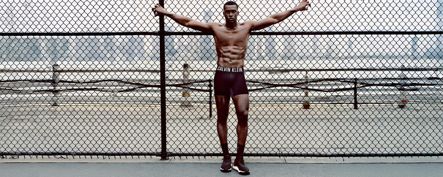 Mann in schwarzer Calvin Klein Unterwäsche, stehend mit ausgebreiteten Armen vor Maschendrahtzaun im Freien.