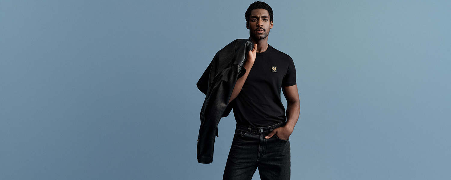 Mann in schwarzem T-Shirt von Belstaff mit Logo und schwarzer Jeans, schwarze Lederjacke über der Schulter vor blauem Hintergrund.