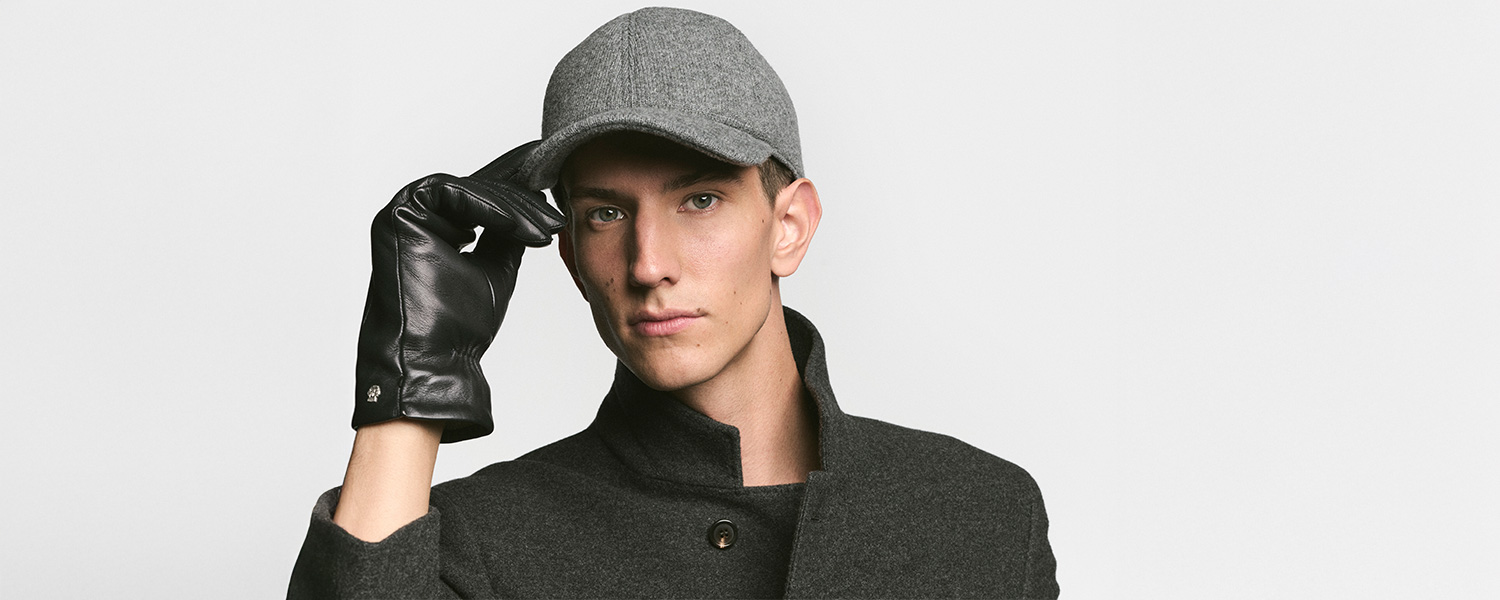 Ein Mann trägt Roeckl Accessoires: elegante schwarze Lederhandschuhe und eine graue Wollcap, kombiniert mit einem dunkelgrauen Mantel für einen stilvollen, urbanen Look.