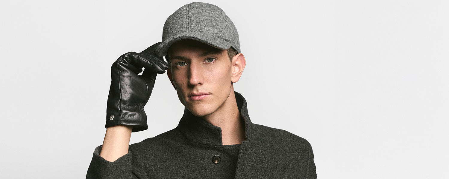 Ein Mann trägt Roeckl Accessoires: elegante schwarze Lederhandschuhe und eine graue Wollcap, kombiniert mit einem dunkelgrauen Mantel für einen stilvollen, urbanen Look.