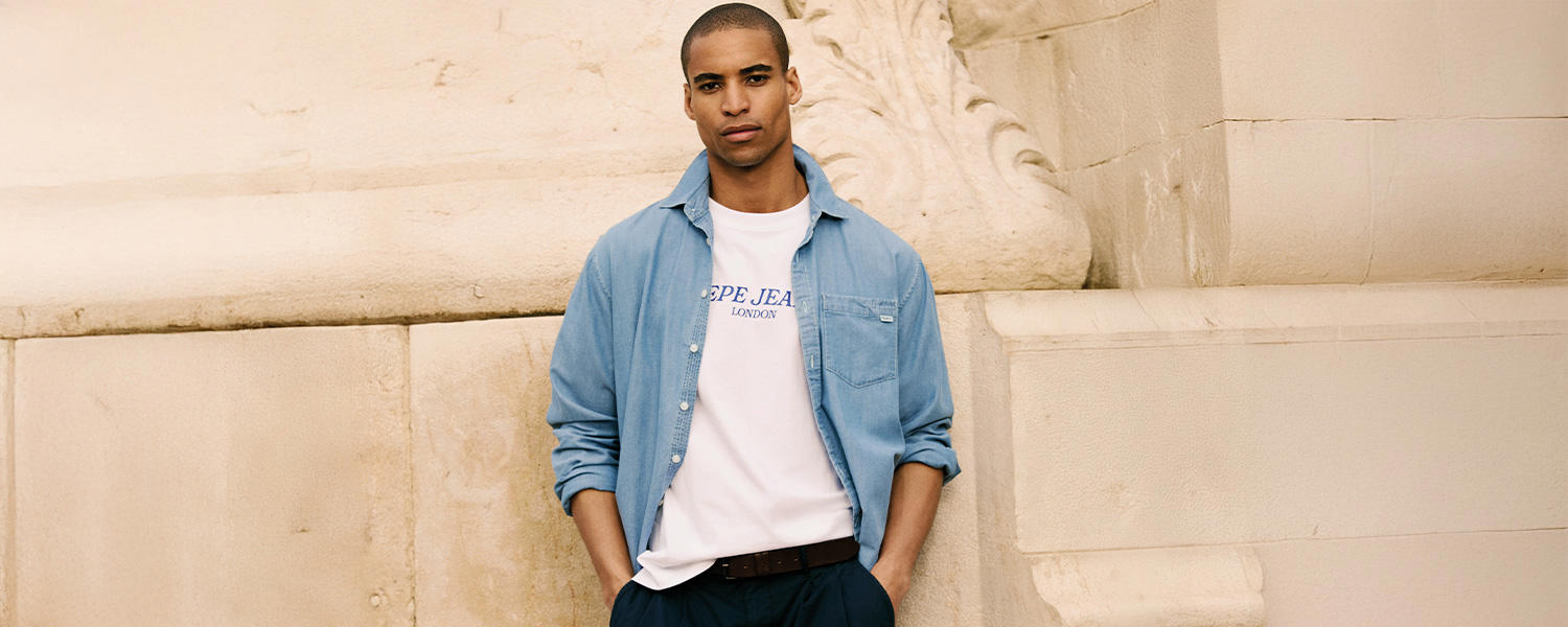 Mann in einem weißen Pepe Jeans T-Shirt mit blauer Aufschrift und offener hellblauer Jeansjacke, lässig an einer sandfarbenen Steinwand lehnend.