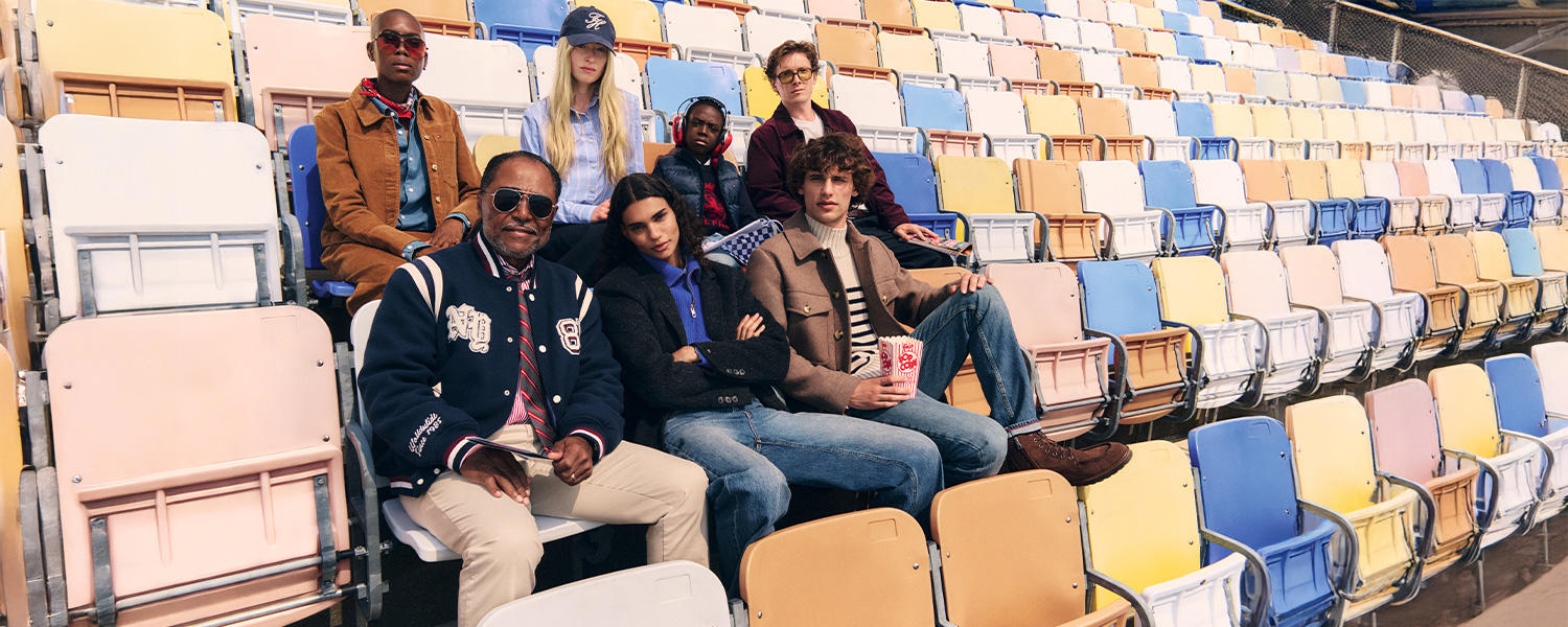 Eine lebendige Kampagnenszene von Tommy Hilfiger: Eine diverse Gruppe sitzt in bunten Stadion-Sitzen, gekleidet in typische Tommy-Hilfiger-Mode mit sportlich-preppy Einflüssen.