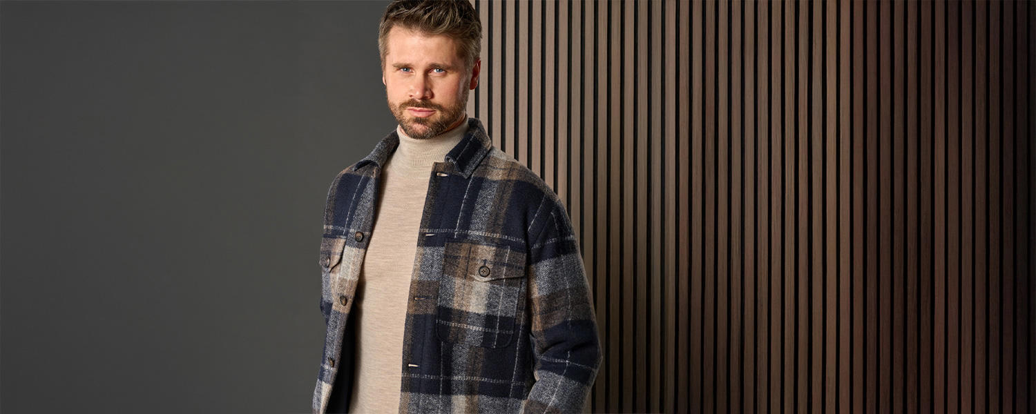 Ein modernes Markenmotiv von Venti: Ein Mann trägt ein kariertes Overshirt in Blau- und Brauntönen über einem hellen Rollkragenpullover. Der Hintergrund mit Holzpaneelen unterstreicht die Kombination aus urbanem Chic und klassischer Eleganz.