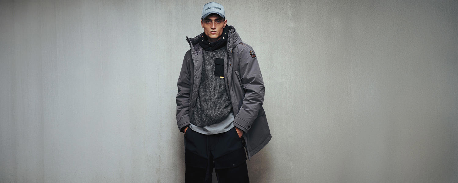 Mann in grauer Winterjacke, grauem Pullover und Cap von Parajumpers vor grauer Betonwand, lässig mit den Händen in den Taschen.