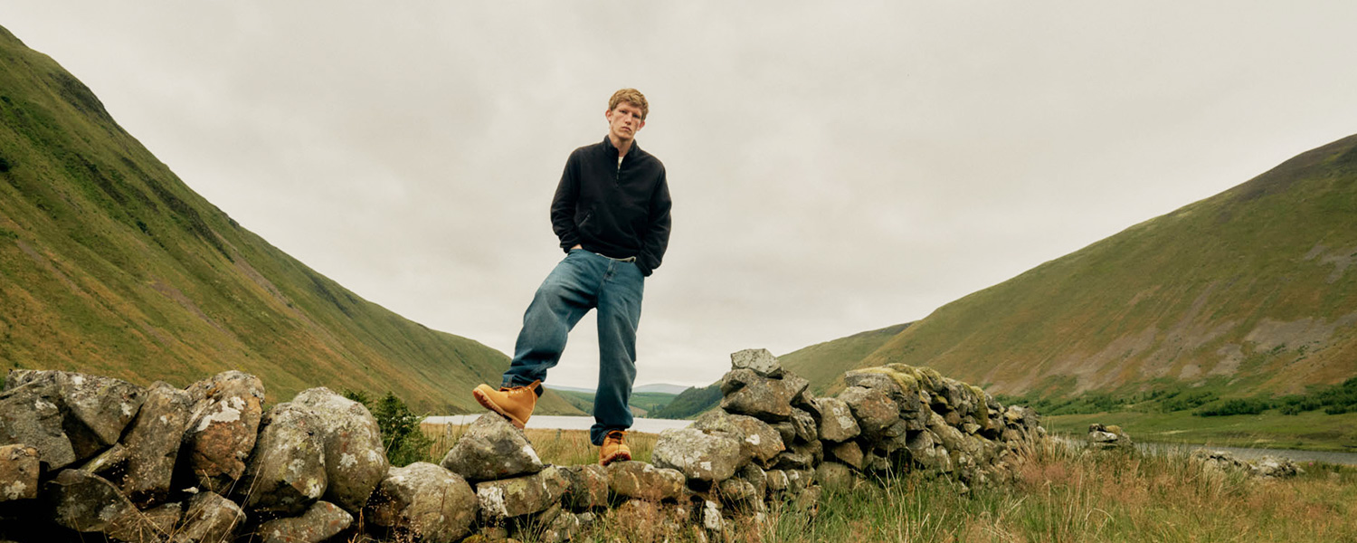 Ein authentisches Outdoor-Motiv von Timberland: Ein Mann trägt robuste Timberland-Boots und Jeans, während er auf einer Steinmauer inmitten einer weiten, grünen Landschaft steht – Sinnbild für Abenteuer und Naturverbundenheit.