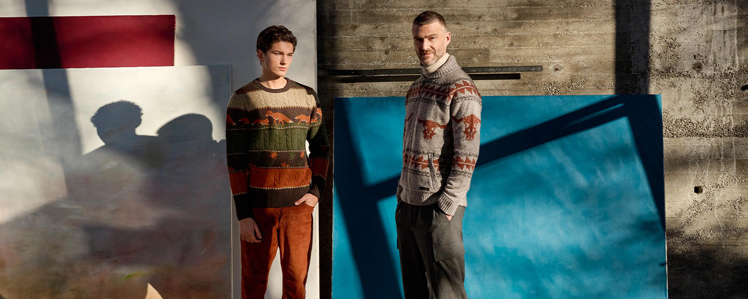 Zwei Männer in Ragman-Pullovern mit auffälligen Mustern: links ein mehrfarbiger Pullover in Grün-, Braun- und Orangetönen, rechts ein grauer Rollkragenpullover mit roten Musterdetails, posierend in einem künstlerischen Setting mit gemalten Hintergründen.