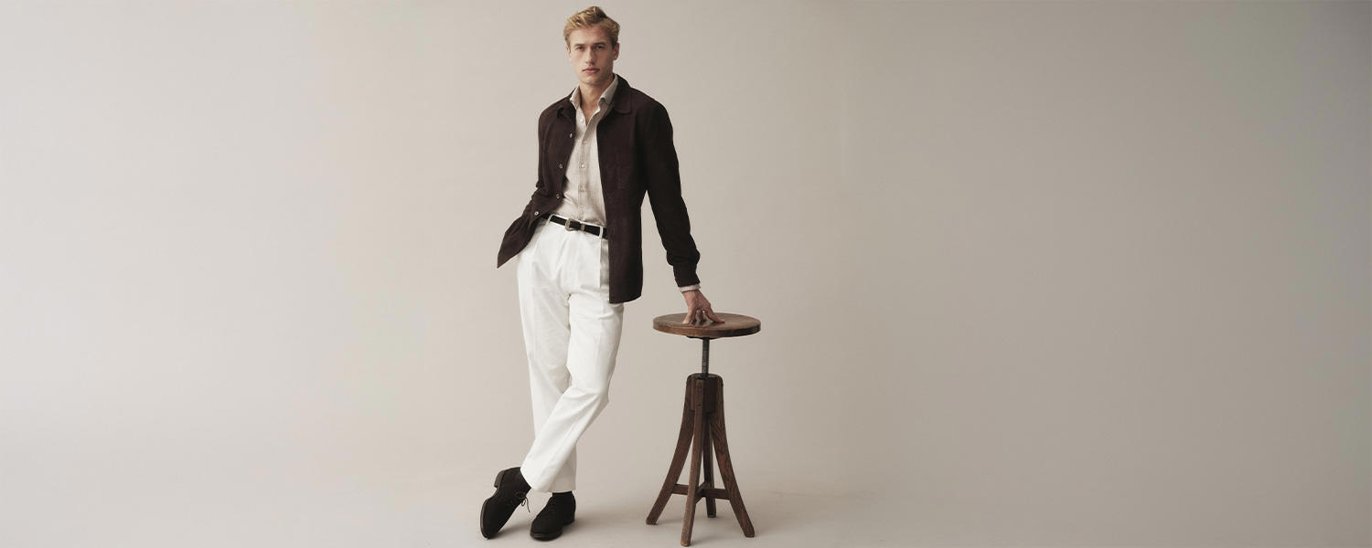 Ein elegantes Kampagnenmotiv von Stenströms: Ein Mann trägt ein beiges Hemd mit brauner Jacke, kombiniert mit weißen Hosen und schwarzen Schuhen, lässig angelehnt an einen Hocker vor neutralem Hintergrund.
