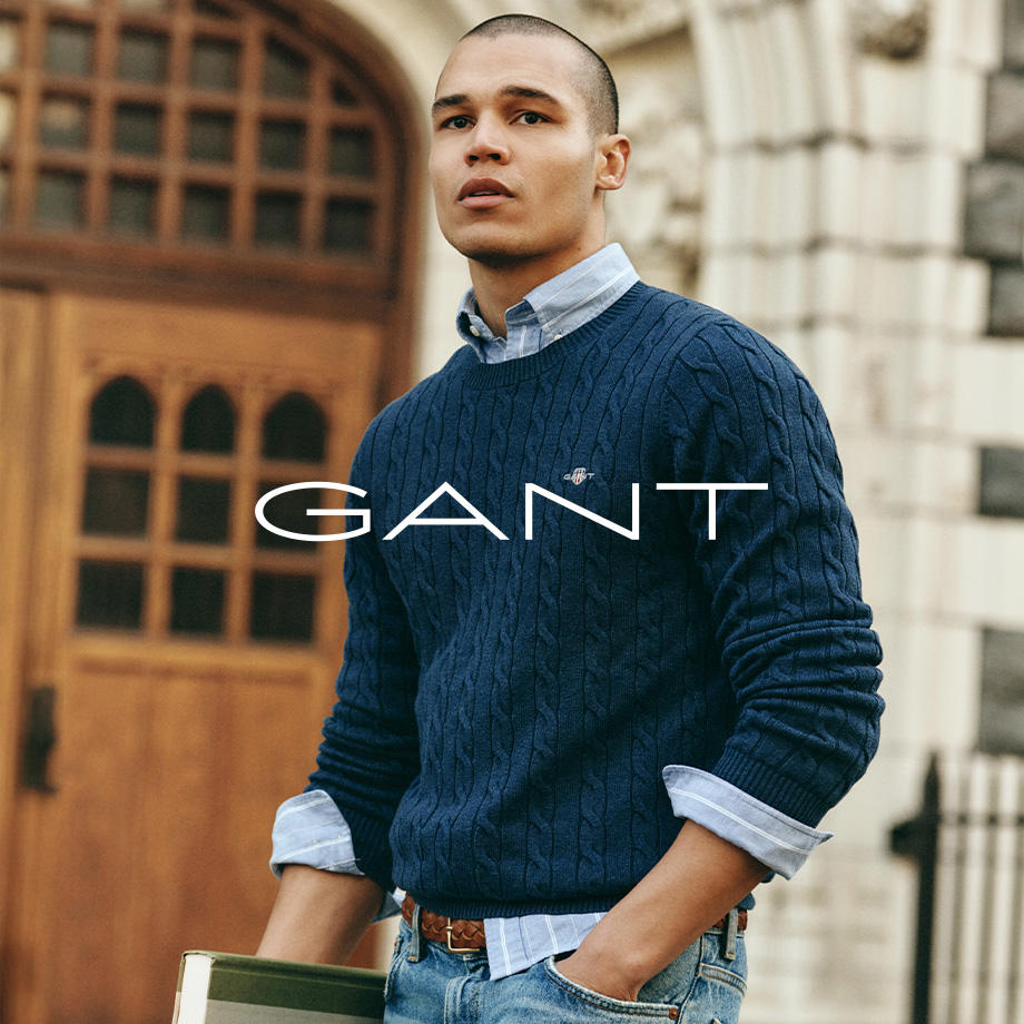 Mann in navyblauem Strickpullover mit Zopfmuster über hellblauem gestreiftem Hemd, kombiniert mit Jeans, stehend vor Gebäude