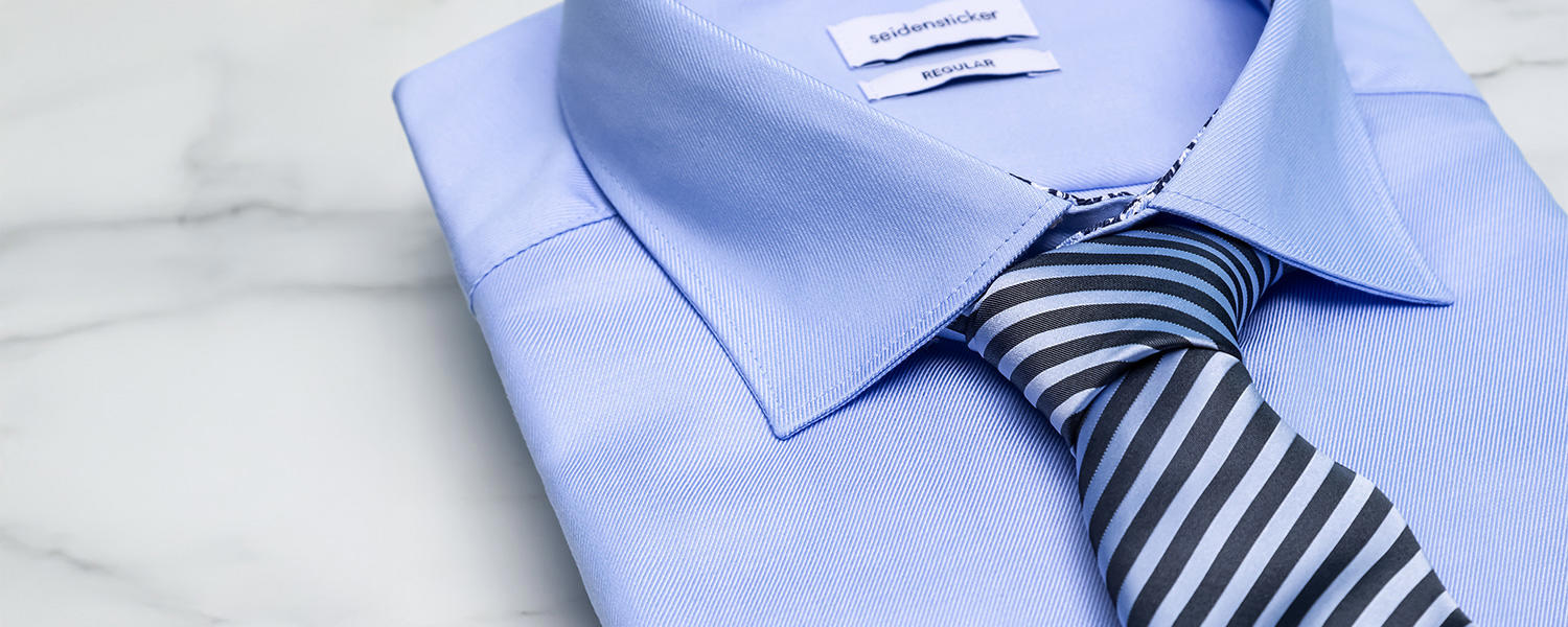 Gefaltetes hellblaues Hemd der Marke Seidensticker mit blau gestreifter Krawatte, liegend auf einem marmorierten, hellen Untergrund.