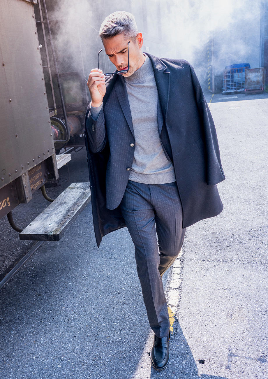 Ein Mann in einem grau gestreiften Anzug mit hellgrauem Rollkragenpullover geht vor einer alten Eisenbahn entlang. In einer Hand hält er eine Brille, die er sich gerade abzieht. Im Hintergrund liegt leichter Nebel und die Lichtstimmung ist kühl und gedämpft.