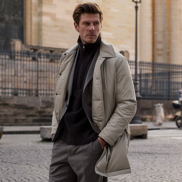 Grauer Wollmantel über Strick und Blazer, kombiniert mit Anzughose, urbaner Business-Casual Look.