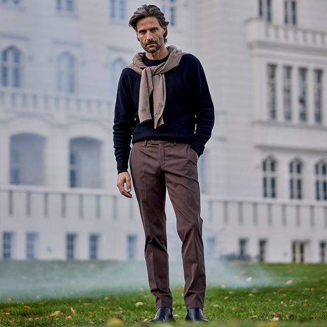 Dunkler Pullover mit beiger Strickjacke über den Schultern, dazu braune Chinos und Chelsea-Boots, entspannter Casual-Style.