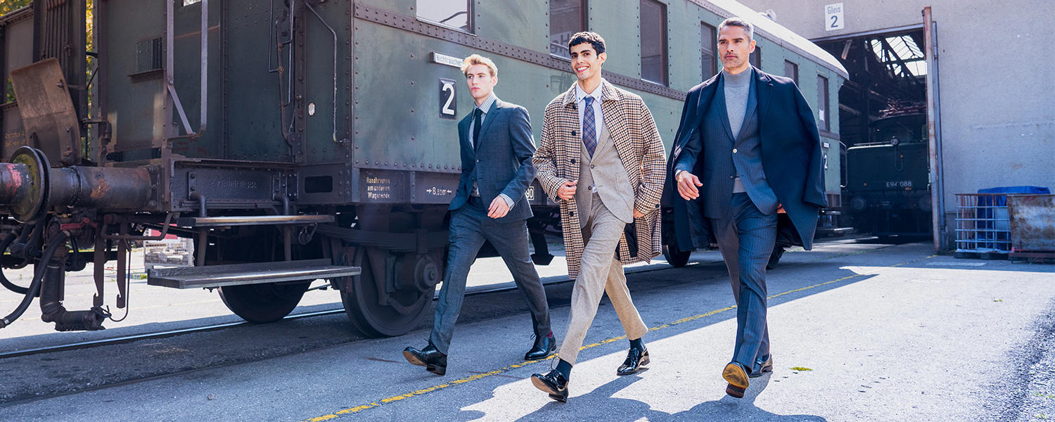 Drei Männer laufen an einem Zugabteil entlang. Der links außen trägt einen karieren Mantel, ein gestreiftes Hemd und eine dunkle Krawatte. Der in der Mitte trägt einen Karomantel, einen beigen Anzug, ein weißes Hemd und eine karierte Krawatte. Der Mann rechts trägt einen Mantel, einen gestreiften Anzug und einen grauen Rollkragenpullover.