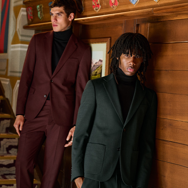 Zwei Männer in eleganten Anzügen, dunkelgrün und bordeaux, kombiniert mit schwarzen Rollkragenpullovern, moderner Business-Look.