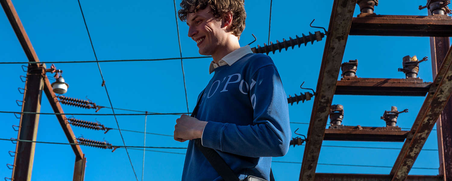 Mann im blauen Logo-Sweatshirt über weißem Hemd, entspannter Casual-Look unter freiem Himmel.