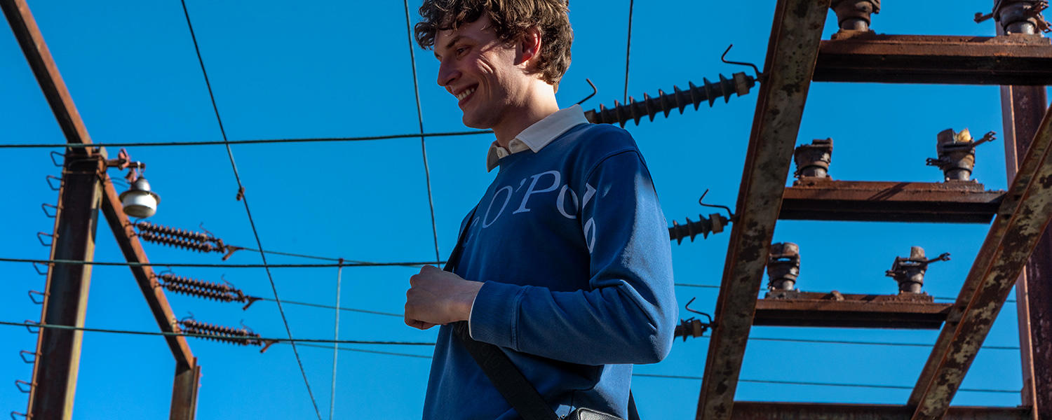 Mann im blauen Logo-Sweatshirt über weißem Hemd, entspannter Casual-Look unter freiem Himmel.