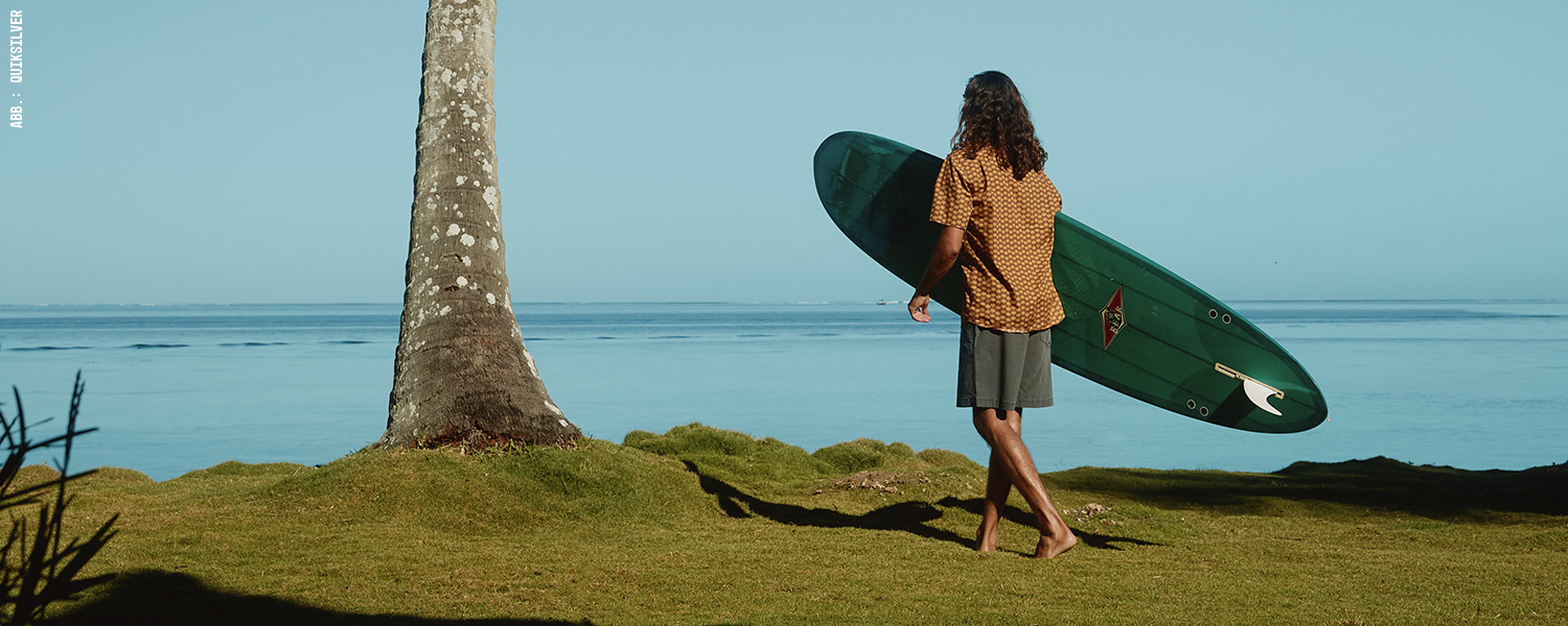 Mann mit Surfboard am Strand – cooler Freizeitlook mit lockerer Badeshorts.