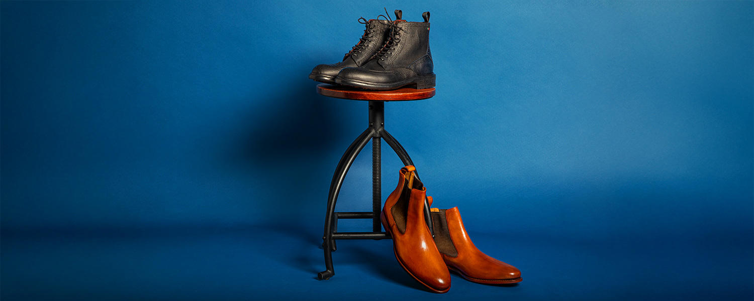 Schwarze Schnürboots aus Leder im robusten Design, platziert auf einem Hocker und elegante cognacfarbene Chelsea Boots aus Glattleder auf dem Boden stehend vor blauem Hintergrund.