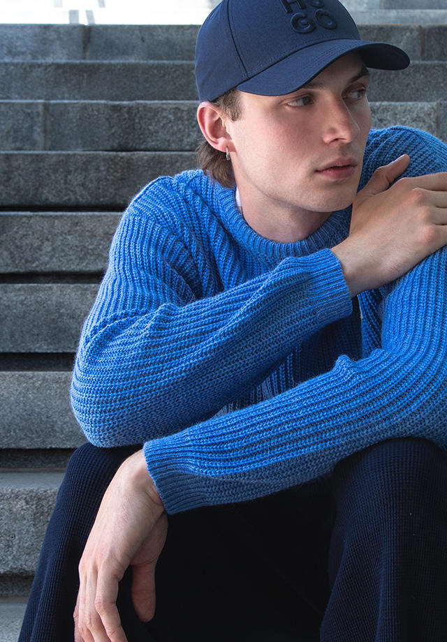 Blauer Strickpullover im lockeren Casual-Look, kombiniert mit Basecap und dunkler Hose.