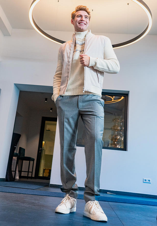 Älterer Mann in grauer Jacke mit beigem Zopfmuster-Rollkragenpullover und heller Hose, sportlich mit Sneakers