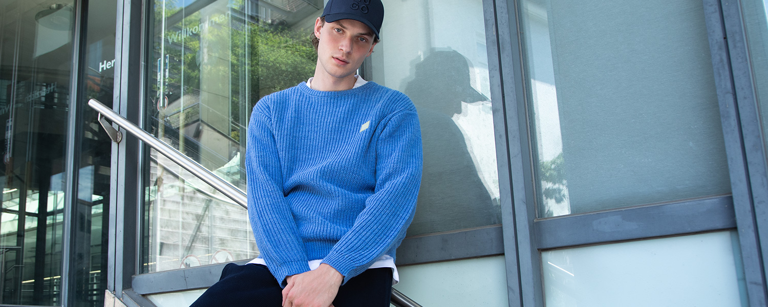 Lässiger Streetstyle mit blauem Strickpullover, weißem Shirt und Cap – moderner Look mit sportivem Flair.