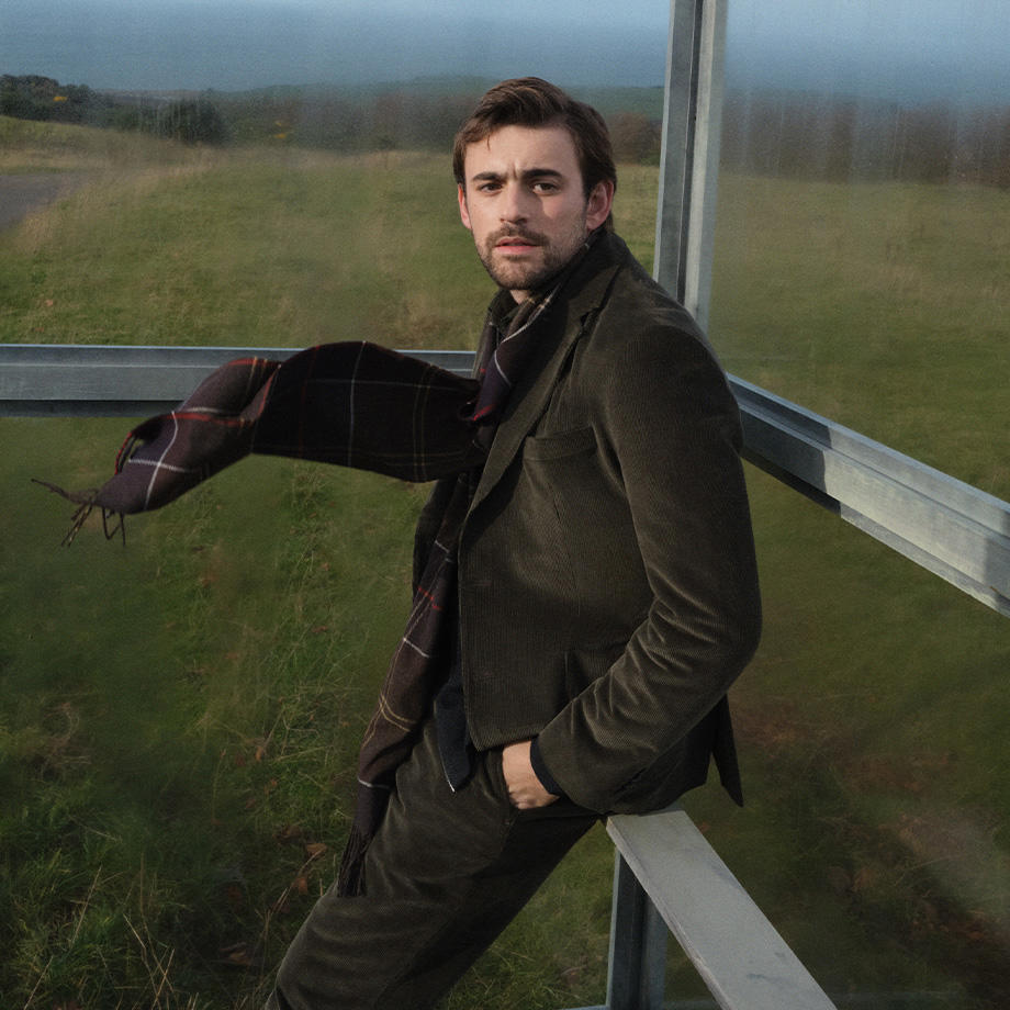 Mann in navyblauer Barbour-Jacke, grauem Schal und Fischerhut lehnt lässig an einem Land Rover – authentischer Outdoor-Look aus der Barbour Tartan Kollektion.