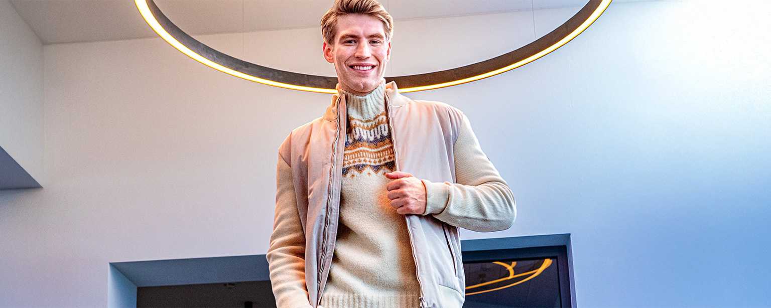 Lächelnder Mann in beigem Norwegerpullover und hellem Blouson unter modernem Lichtring – gemütliche Strickmode für stilvolle Winterlooks bei Herrenausstatter.de.