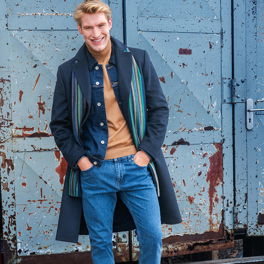 Mann in blauem Mantel, Jeans und beigefarbenem Pullover mit Schal lehnt an alter Metalltür.