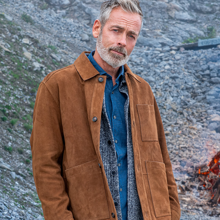 Mann in brauner Wildlederjacke mit Denim-Hemd steht vor felsiger Berglandschaft.